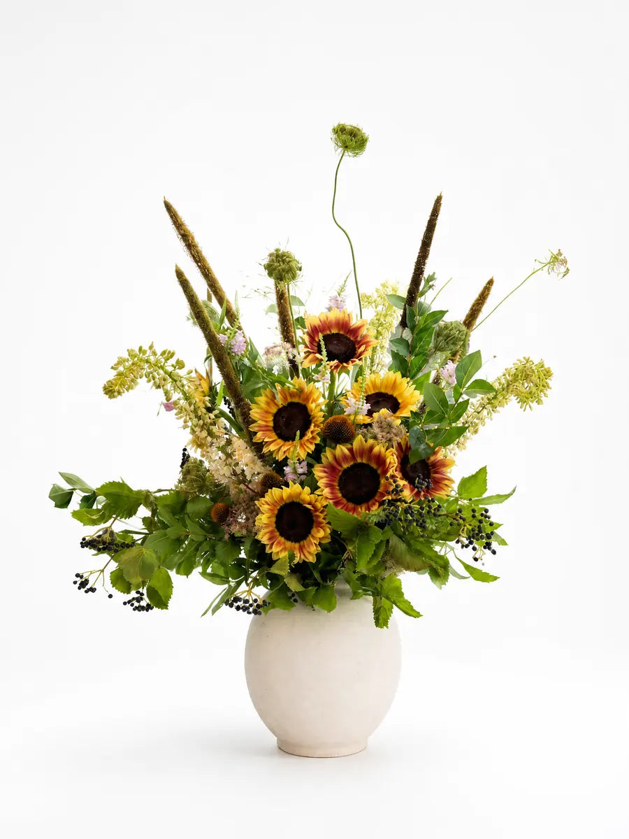 Yellow and rust-colored sunflowers, berries, and leafy branches in a cream ceramic vase