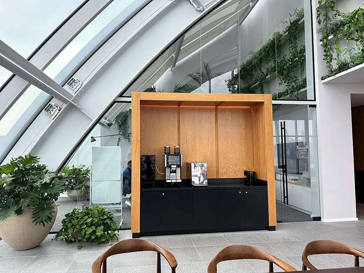 A timber coffee kiosk beneath a glazed atrium roof, framed by potted palms and trailing greenery